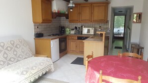 Een compacte keuken met houten kastjes, een koelkast, een magnetron en een spoelbak. Er staat een eettafel met stoelen en er ligt een decoratief plaid op de bank.
