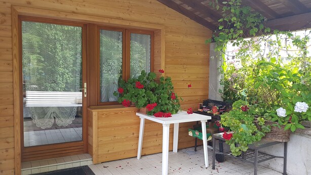Een houten chalet met een veranda, een witte tafel en planten in potten met rode bloemen.