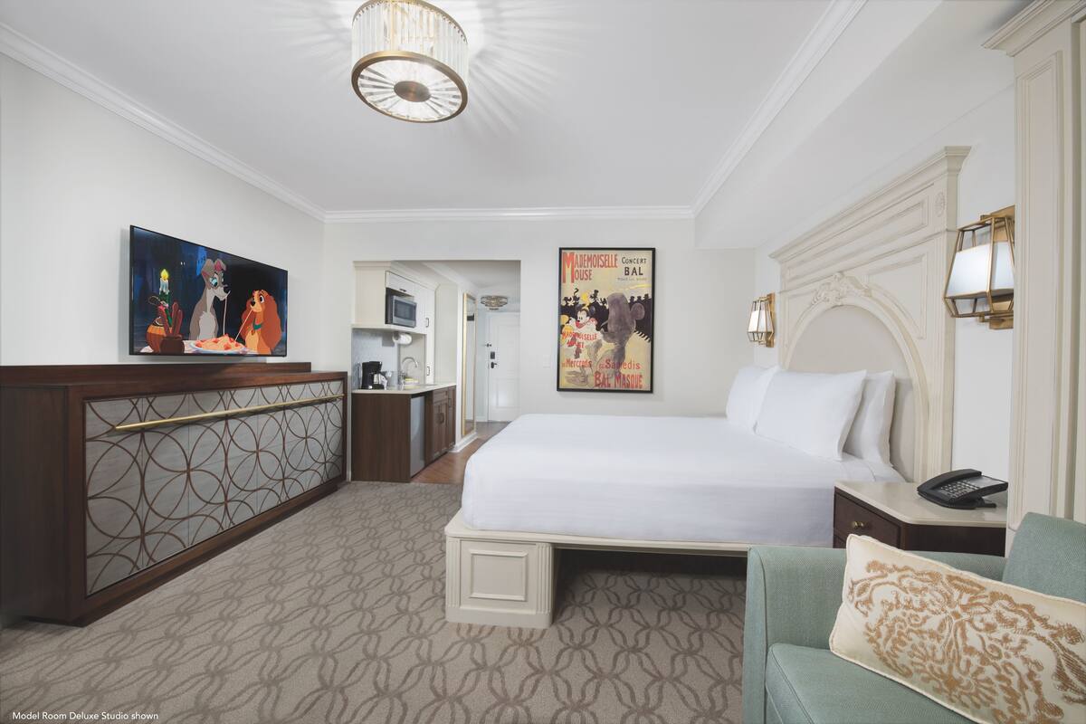 A hotel room with a bed, a desk, a television, a poster, and a chandelier.
