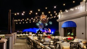 Una terraza en la azotea con mobiliario exterior, vistas a fuegos artificiales y un edificio con toldo azul.
