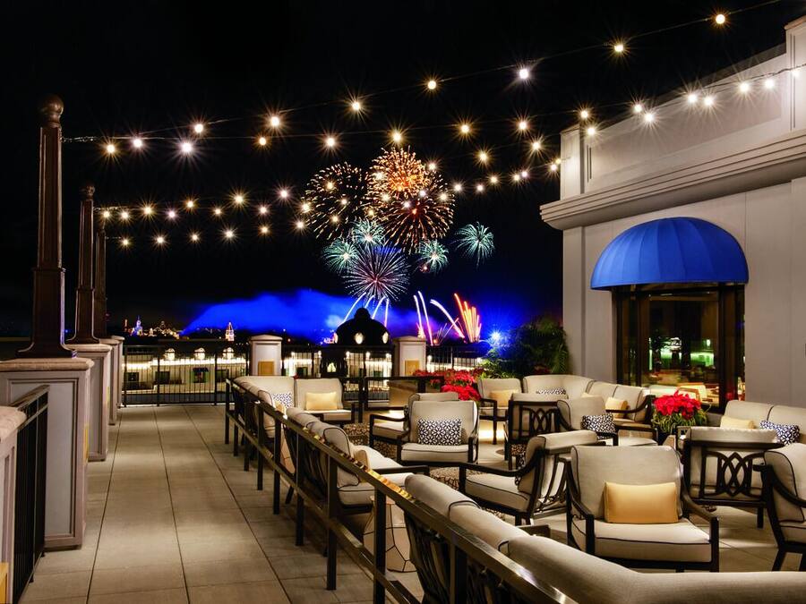 A rooftop terrace with outdoor seating, a view of fireworks, and a building with a blue awning.