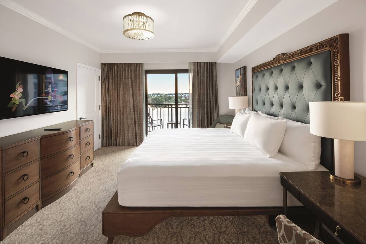 A hotel room with a large bed, a TV on the wall, a desk, and a balcony with a view.