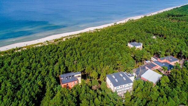 An aerial view of a coastal residential area with dense forest, a beach, and several buildings with flat roofs. - Millennium Spa (Dziwnów)