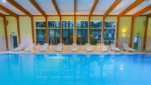 An indoor swimming pool with clear blue water, surrounded by white lounge chairs and large windows offering views of the outside. - Millennium Spa (Dziwnów)