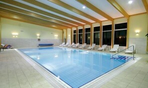 Piscina interna com água azul cristalina, cercada por espreguiçadeiras brancas e piso de azulejos.