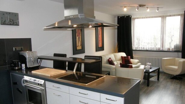 A modern kitchen with a central island, built-in oven, and stainless steel appliances. There is a dining area with a sofa and a coffee table. The room has large windows with curtains. - At the gates of Hamburg in the old country (Mittelnkirchen)