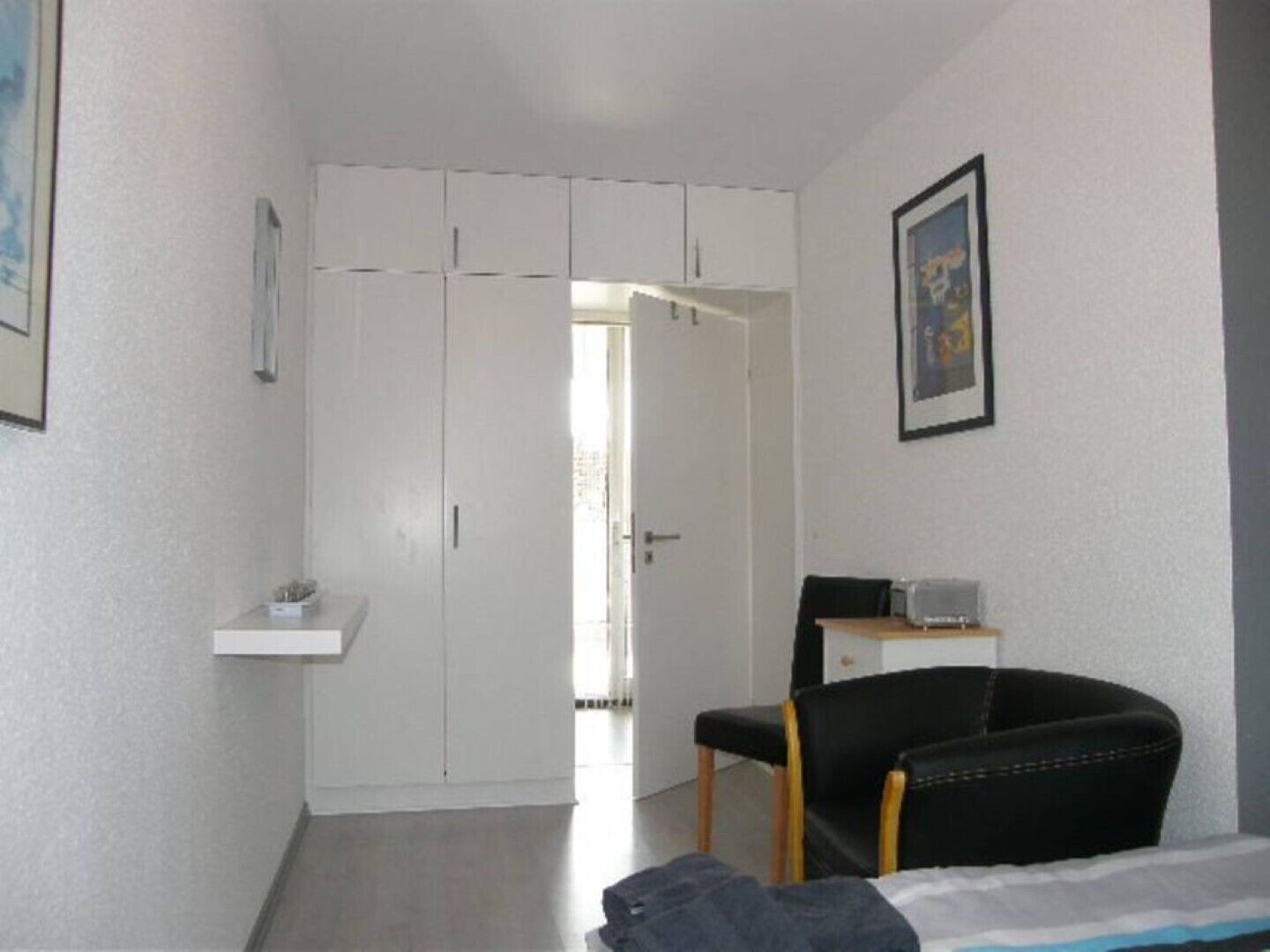 Un couloir étroit avec une armoire blanche, une chaise noire, une petite table avec une radio et un tableau encadré au mur.