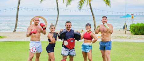 A group of people in athletic wear on a grassy area with a beach and palm trees in the background.