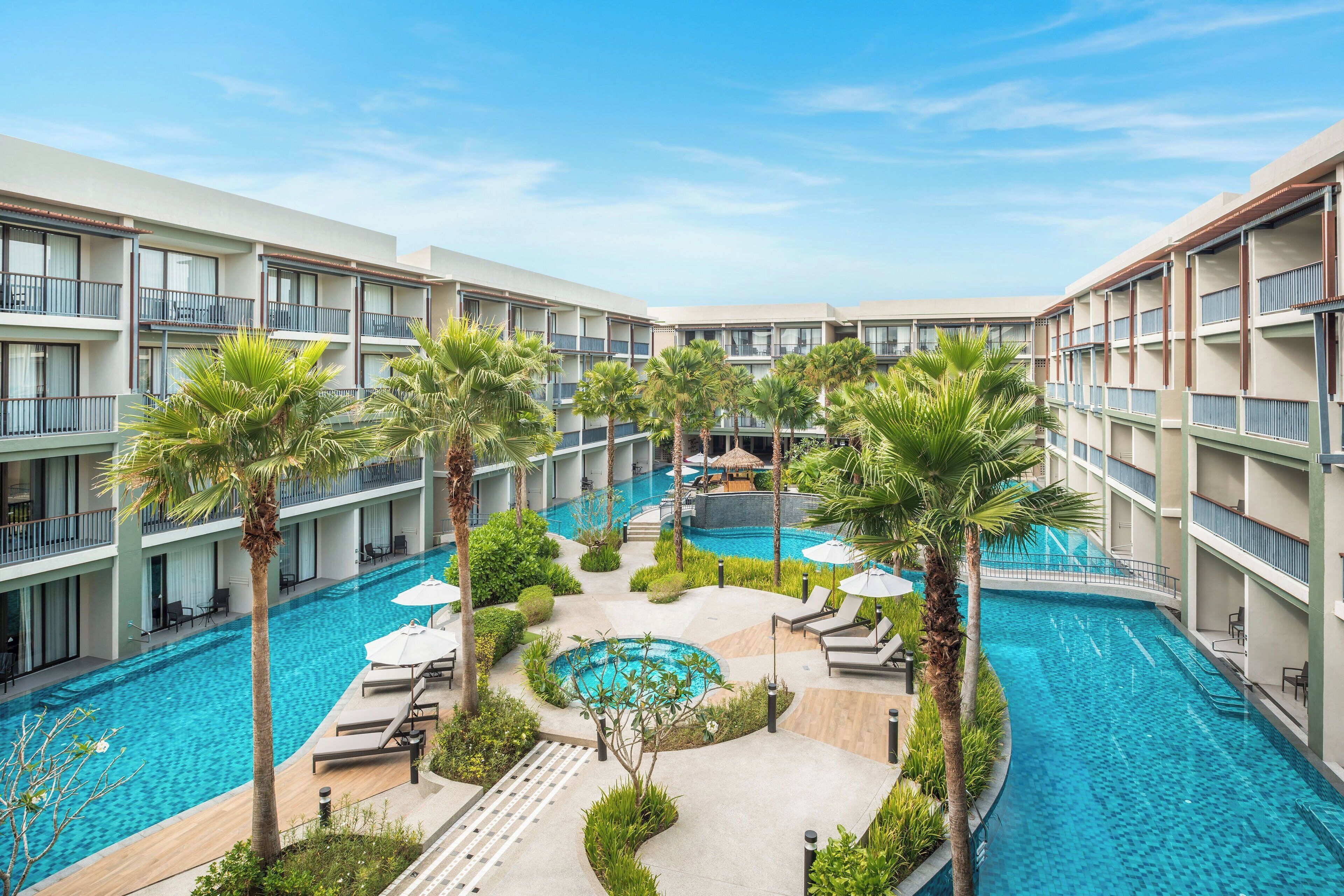 A multi-level hotel with a central swimming pool surrounded by palm trees and lounge chairs.