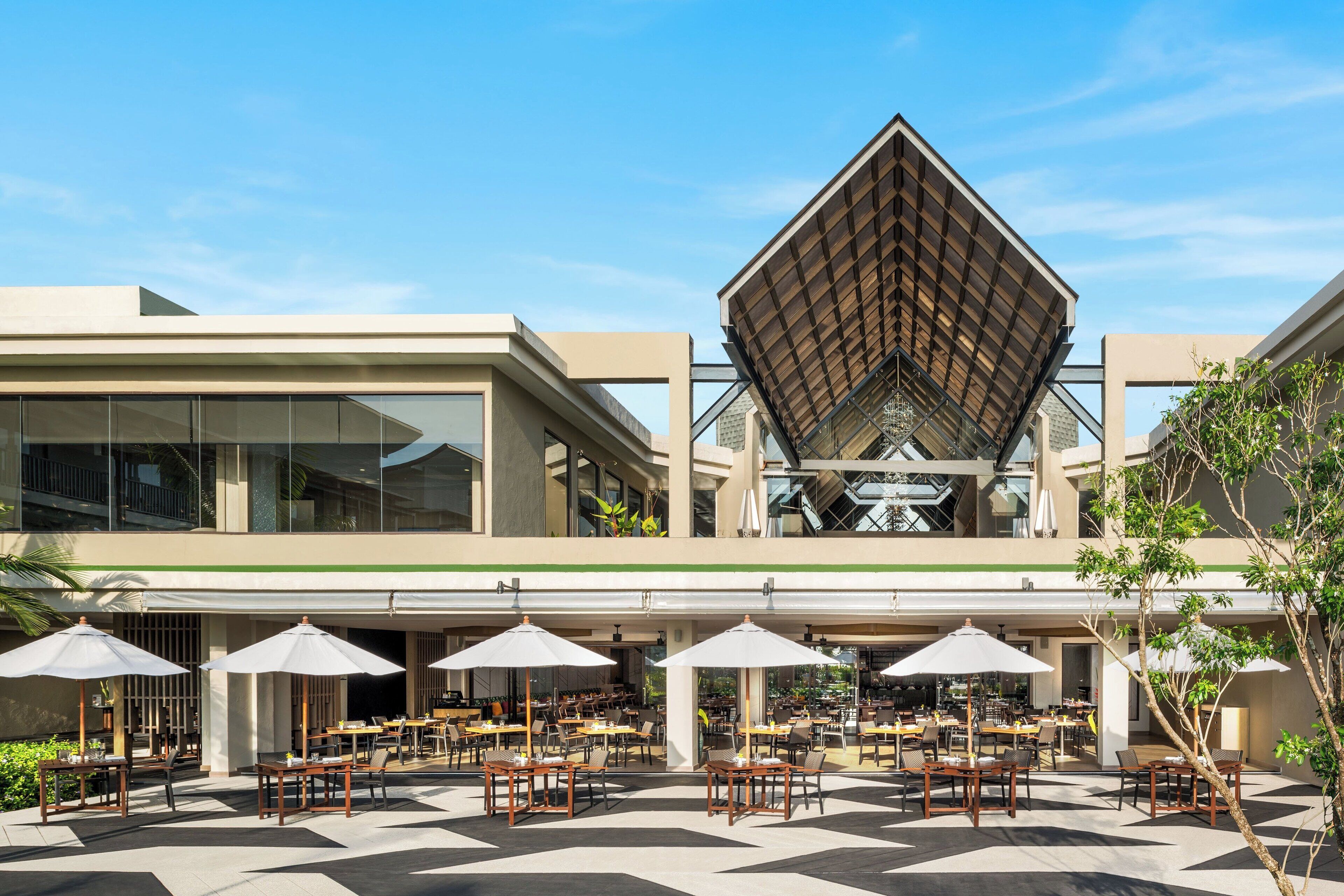 A modern building with a unique geometric roof structure, outdoor seating area with tables and umbrellas, and a mix of indoor and outdoor spaces.