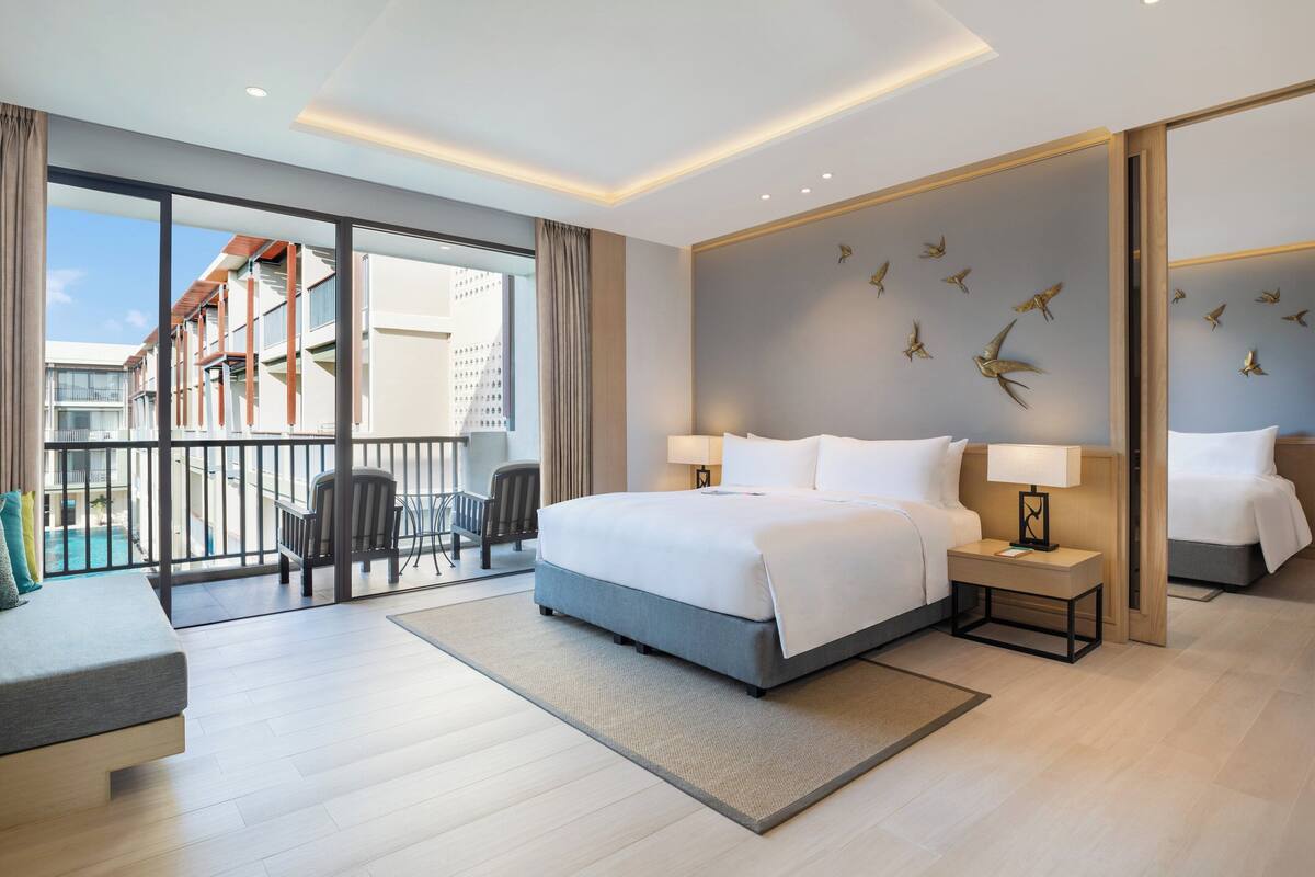 Een moderne hotelkamer met een groot bed, twee nachtkastjes, een zithoek met een fauteuil en een kleintje tafeltje, en een balkon met uitzicht op de stad en een zwembad.