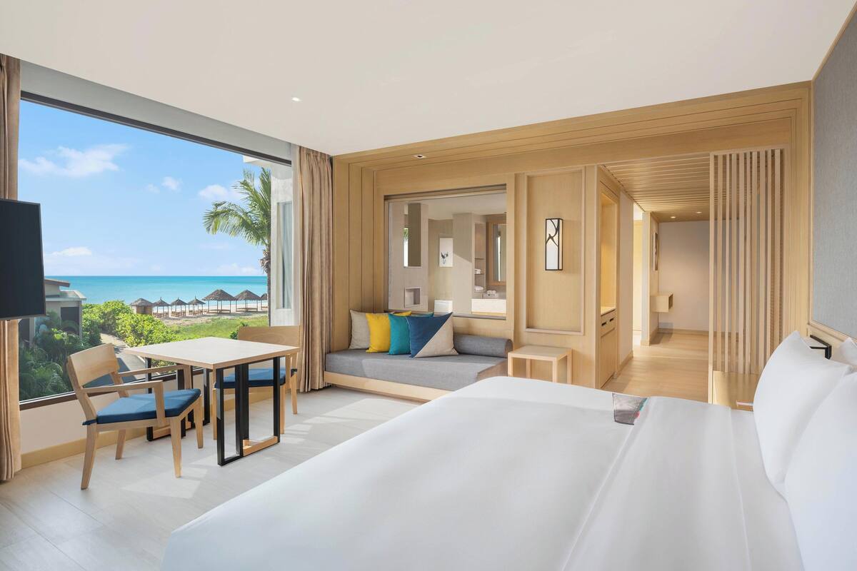 Een moderne hotelkamer met een groot bed, een zithoek met een tafel en stoelen, een bank met kleurrijke kussens, en uitzicht op de oceaan en het strand.