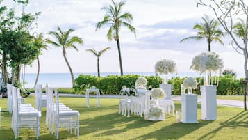 A wedding setup with white chairs and tables, floral arrangements, and palm trees by the ocean.