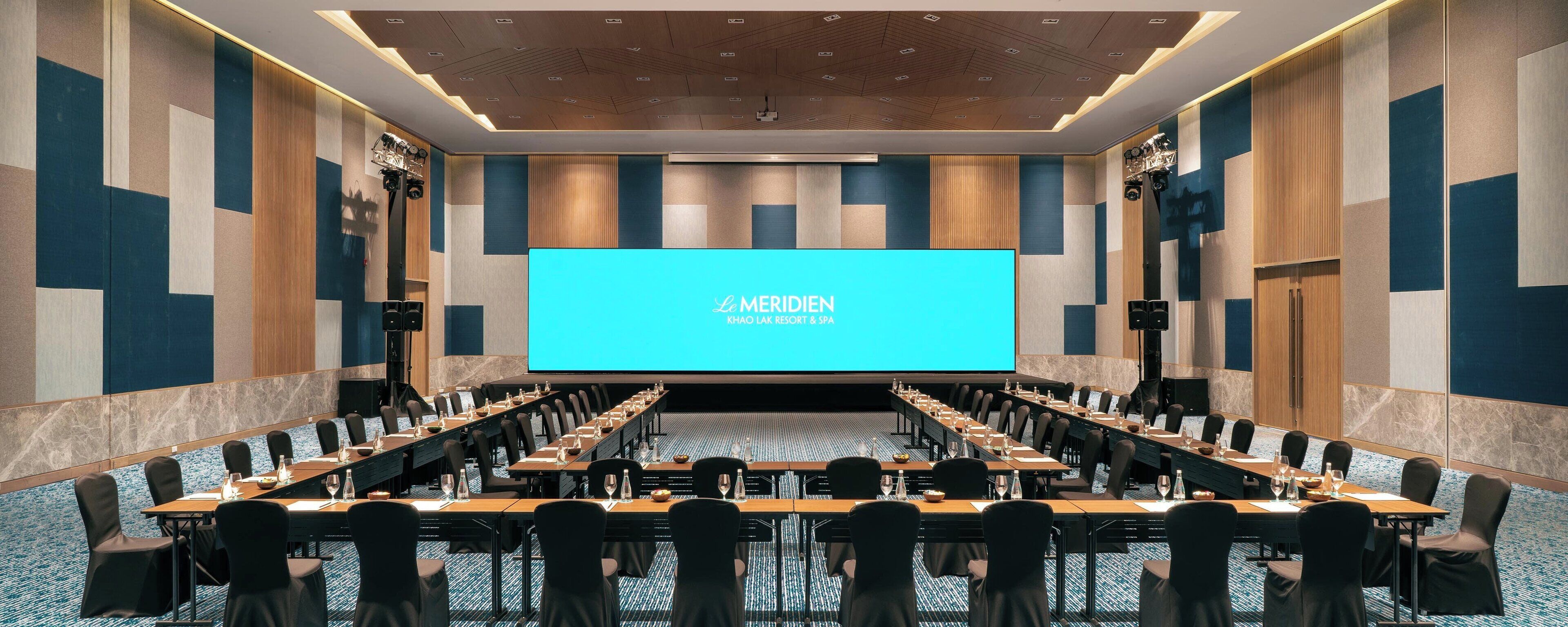Uma sala de conferências com uma grande tela exibindo "Le Meridien Khao Lak Resort & Spa" na frente.