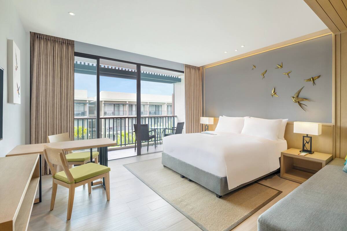 Een moderne hotelkamer met een groot bed, een bureau, stoelen, een balkon met uitzicht op gebouwen en decoratieve muurkunst.
