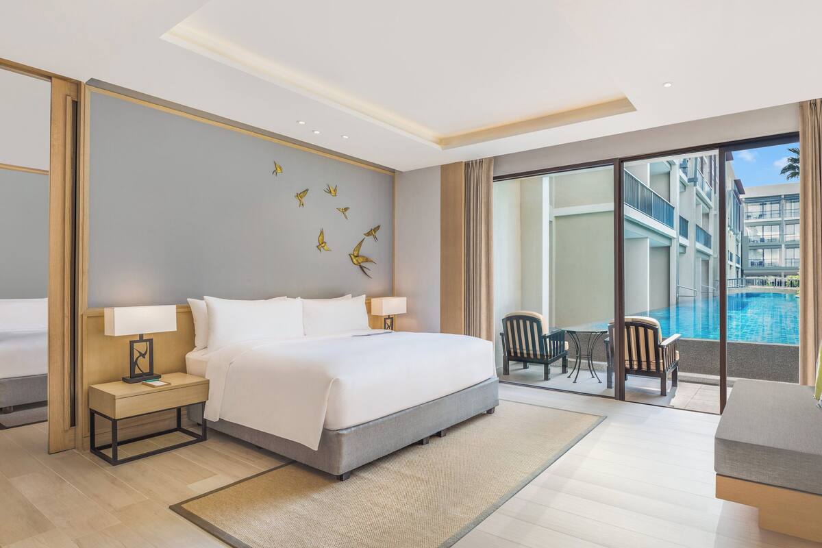 Een hotelkamer met een groot bed, een nachtkastje met een lamp, uitzicht op een zwembad en een balkon met stoelen.