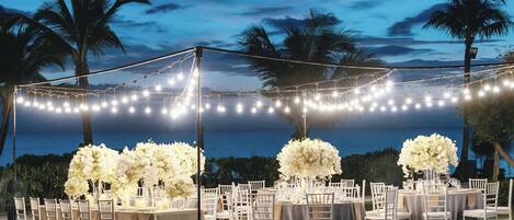 Montaje de boda al aire libre con mesas, sillas y luces colgantes frente a un telón de fondo de palmeras y un atardecer.