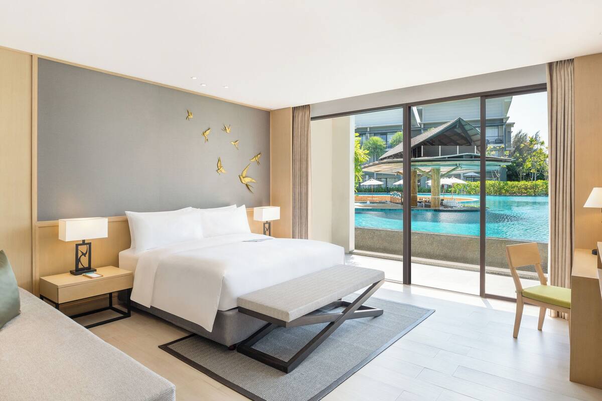Een moderne hotelkamer met een groot bed, een bank, een bureau en uitzicht op een zwembad.