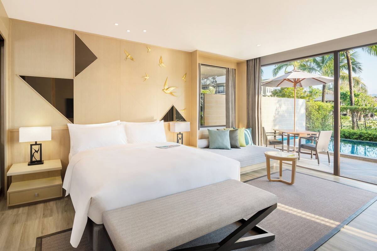 Een moderne hotelkamer met een groot bed, een bank, een kleintje tafeltje en uitzicht op een zwembad.