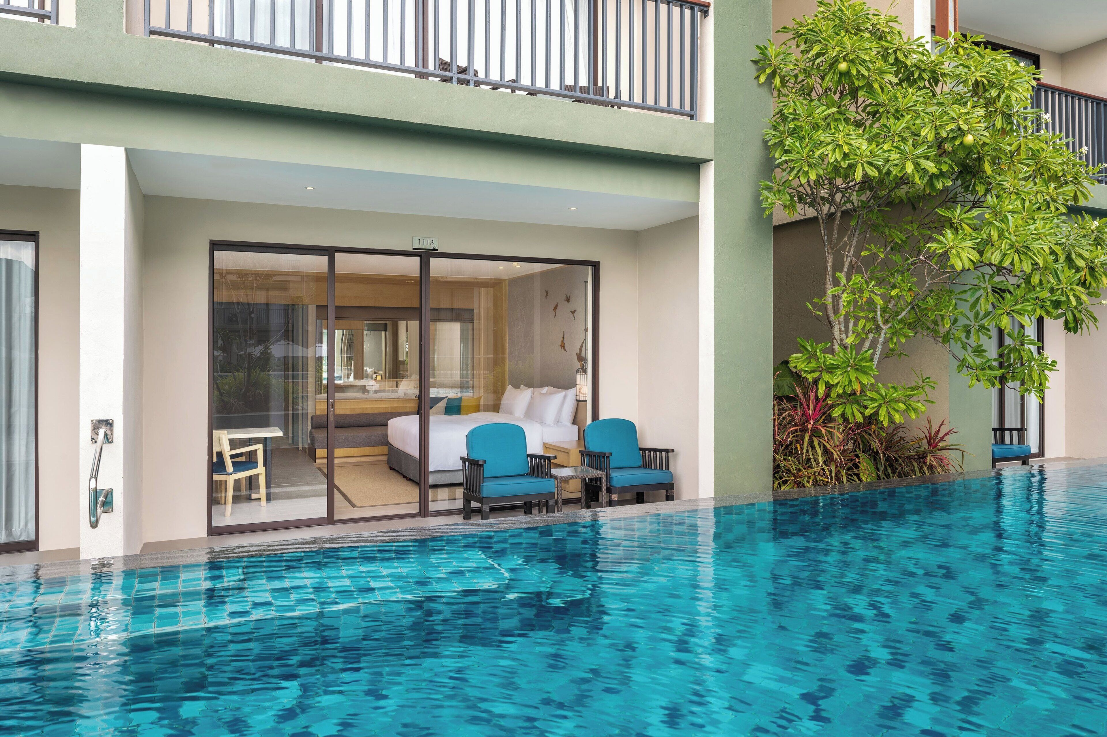 A modern hotel room with a glass door leading to a balcony, a poolside seating area, and a lush green plant.