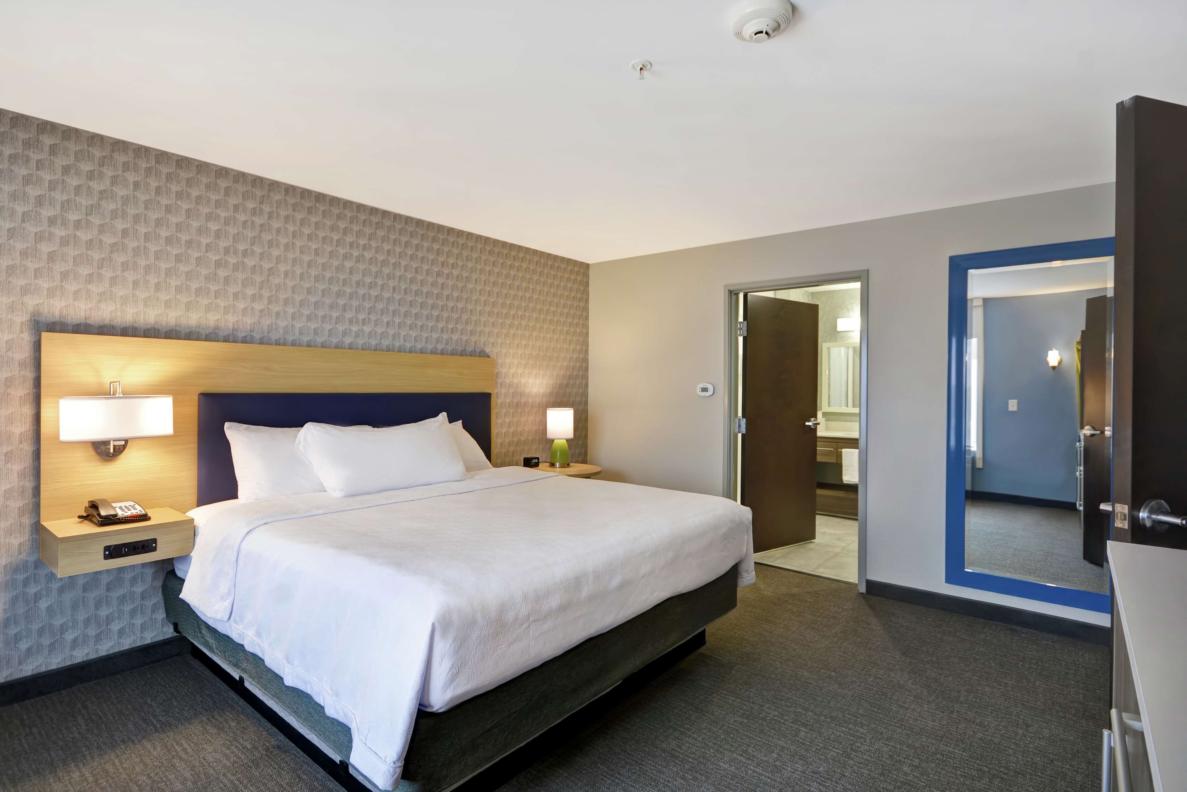 A hotel room with a large bed, bedside lamps, a nightstand, a desk with a lamp, and a door leading to a bathroom.