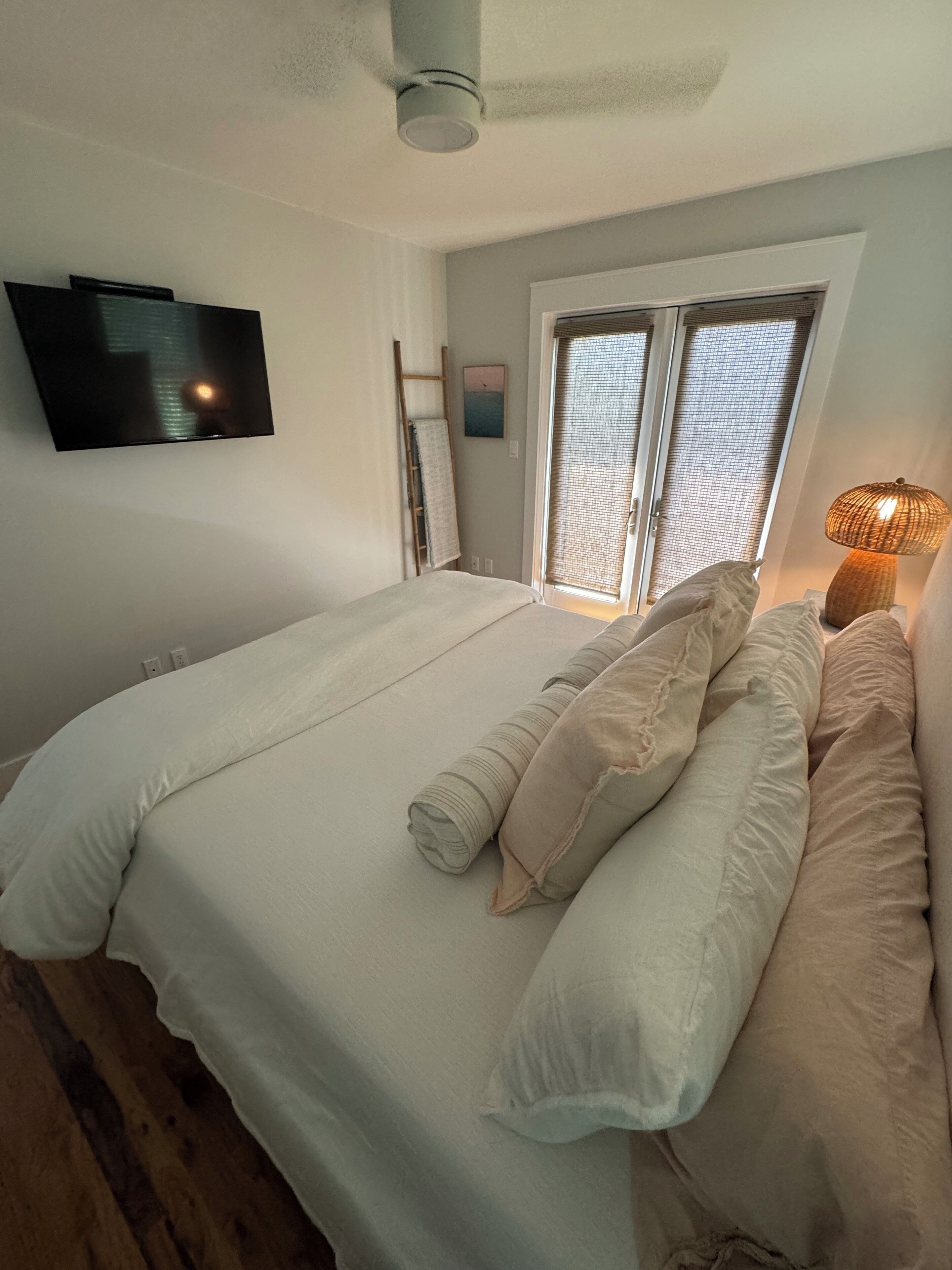 A bedroom with a bed, a TV mounted on the wall, a window with blinds, and a ceiling fan.