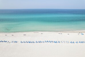 Ein Strand mit weißem Sand, klarem türkisblauem Wasser und Reihen blauer Strandsonnenschirme.