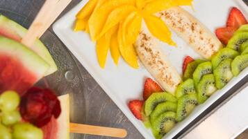 Een wit rechthoekig bord met plakjes mango, kiwi, aardbeien en een banaan op een metalen oppervlak.