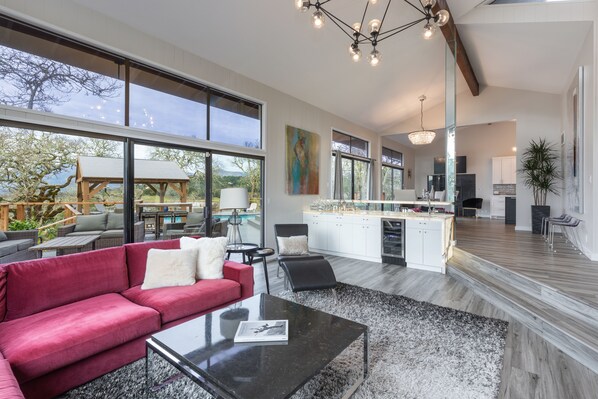 A spacious living area with a red sofa, black coffee table, and large windows offering a view of the outdoors. - Gorgeous Vineyard Views, Large private home w/heated Pool & Hot Tub included! (Glen Ellen)