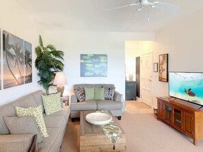 A living room with a grey sofa, a coffee table, a TV on a wooden cabinet, a ceiling fan, and a plant. - Pacific Views w/Private Lanai – Kaha Lani 208 (LIHUE)