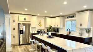 A modern kitchen with white cabinetry, a stainless steel refrigerator, a marble island with bar stools, and a window with blinds. - PONTOON BOAT NOW INCLUDED! 7 BEDS, HOT TUB Updated Lakefront Cottage - Book Now (Norton Shores)