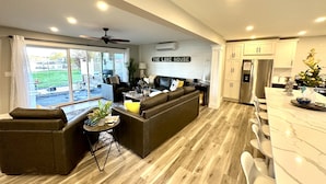 A modern living room with a large L-shaped sectional sofa, a dining area with a marble countertop, and a kitchen with white cabinets and stainless steel appliances. - PONTOON BOAT NOW INCLUDED! 7 BEDS, HOT TUB Updated Lakefront Cottage - Book Now (Norton Shores)