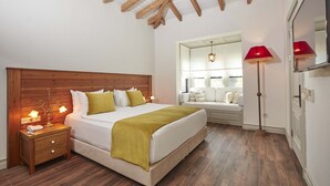 Quarto com cabeceira de madeira, cama com roupa de cama branca e manta verde, mesa de cabeceira com luminárias e um abajur vermelho.