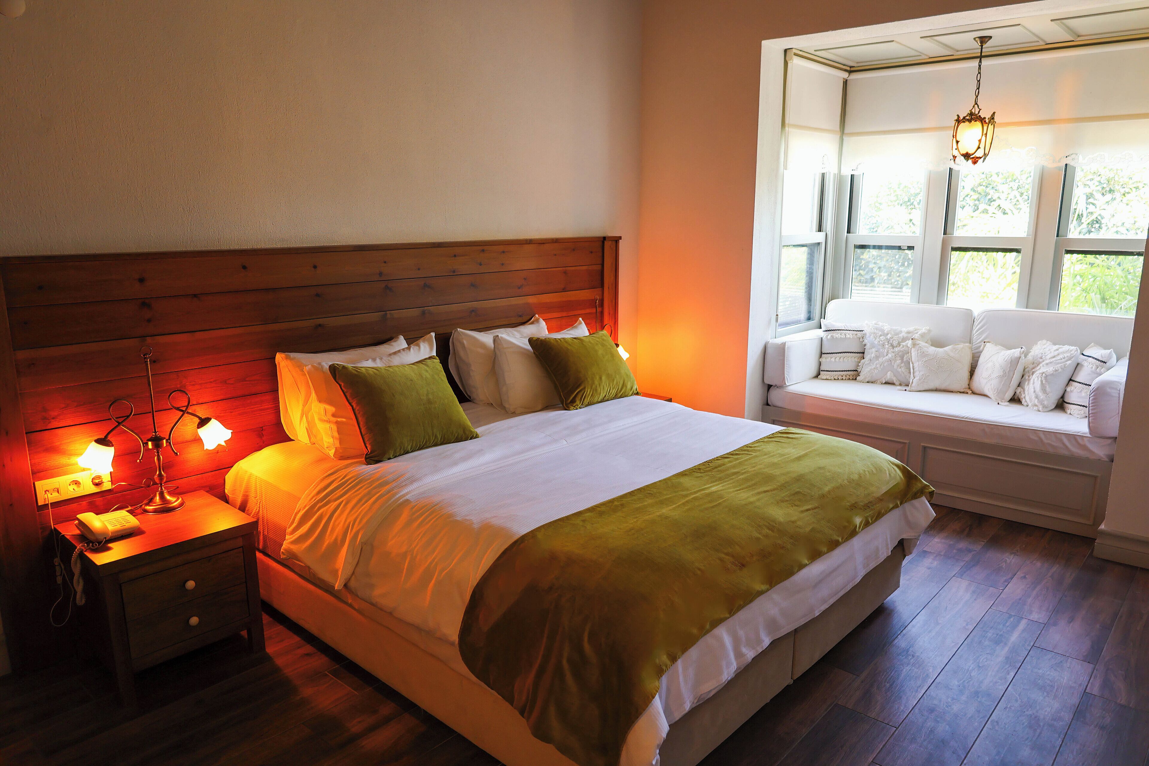 Une chambre à coucher avec une tête de lit en bois, un lit avec une literie blanche et des coussins verts, une table de chevet avec des lampes et un coin fenêtre avec des coussins blancs.
