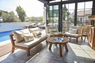 Un área de estar al aire libre iluminada por el sol, con piscina, muebles de madera y una puerta de vidrio que da a un salón junto a la piscina.