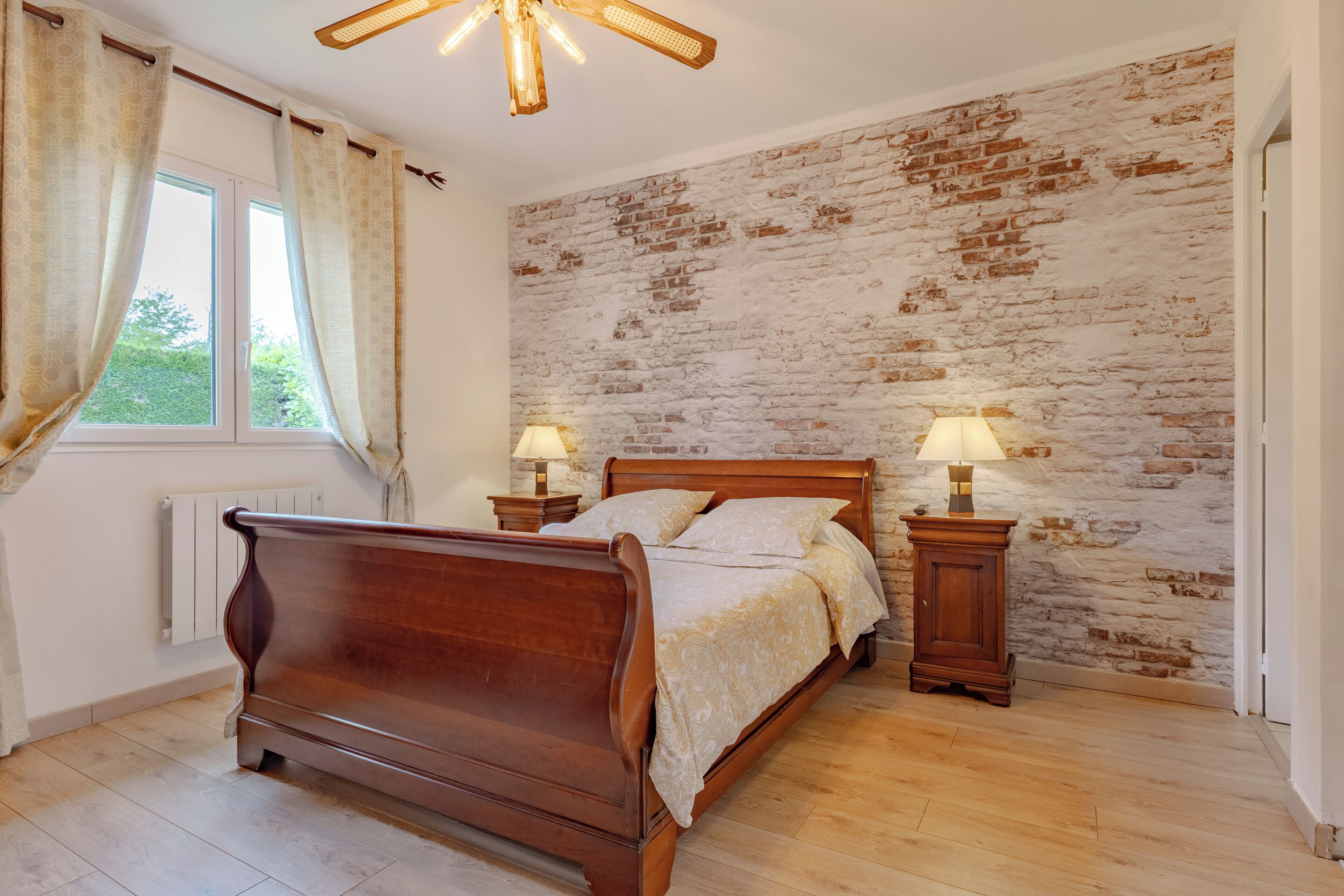 A bedroom with a wooden bed, two bedside lamps, a brick-patterned wall, and a window with curtains.