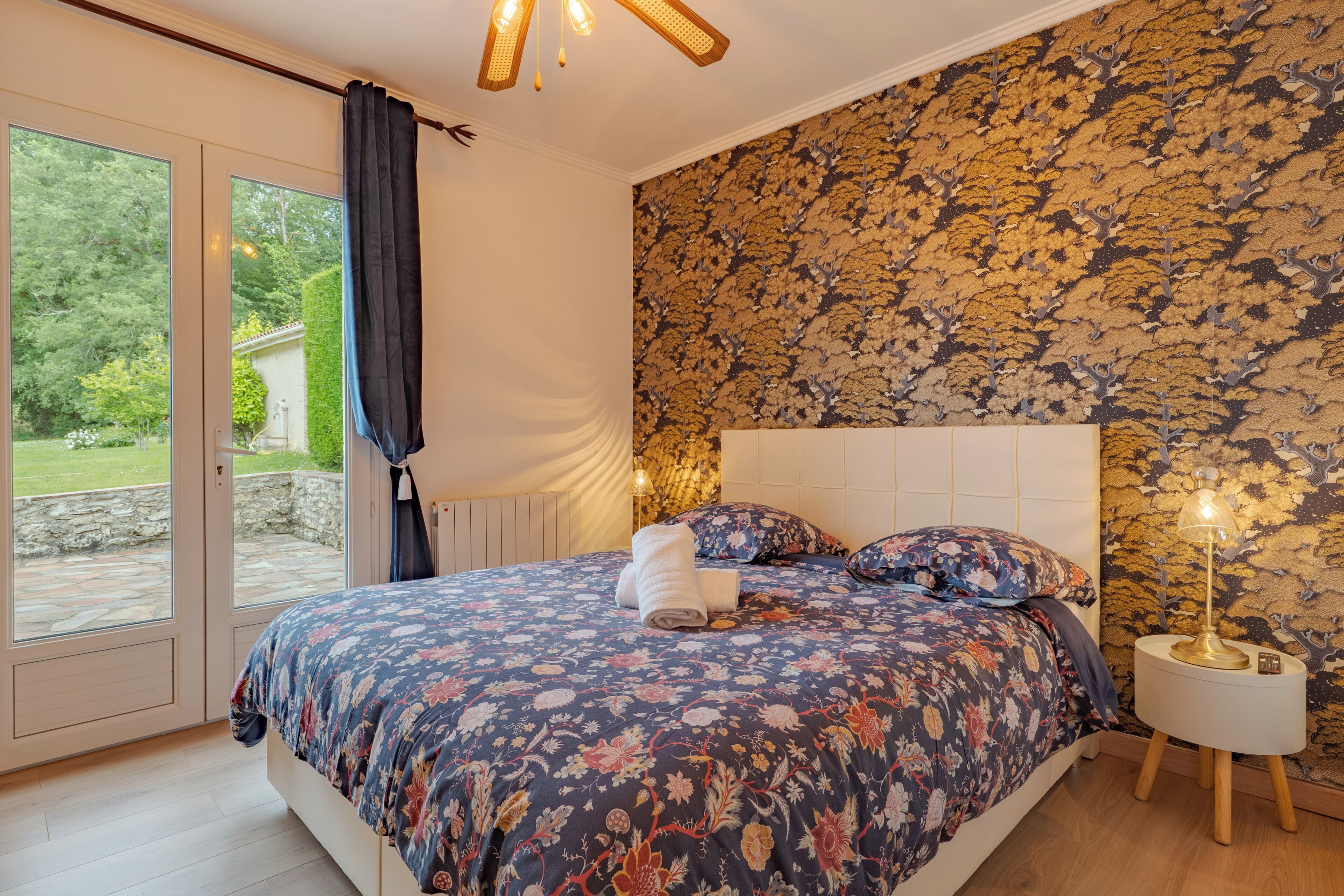 A bedroom with a bed, a nightstand, a lamp, a window with curtains, and a patterned wall.
