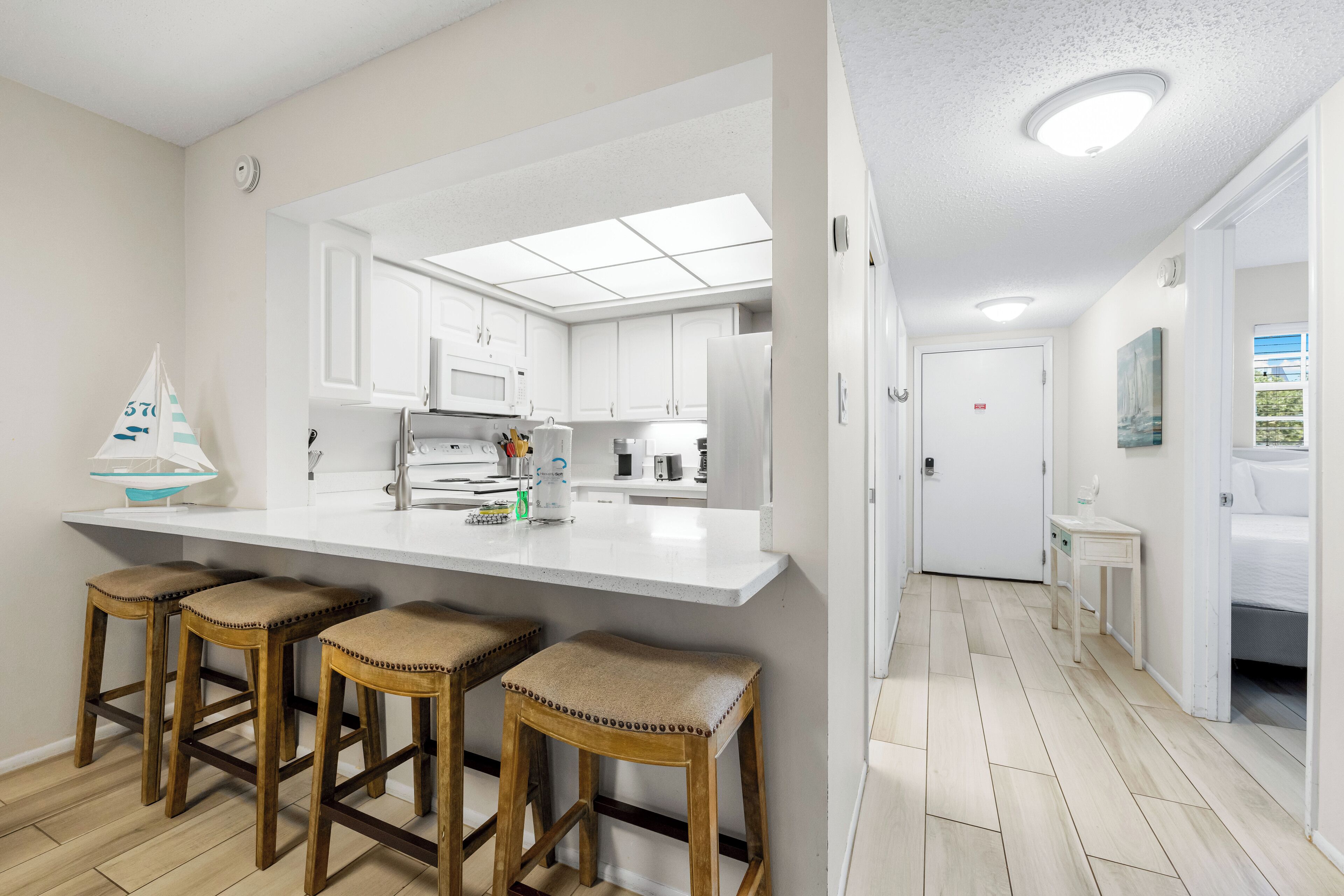 A modern kitchen with white cabinets, a central island with stools, and a view into a bedroom.