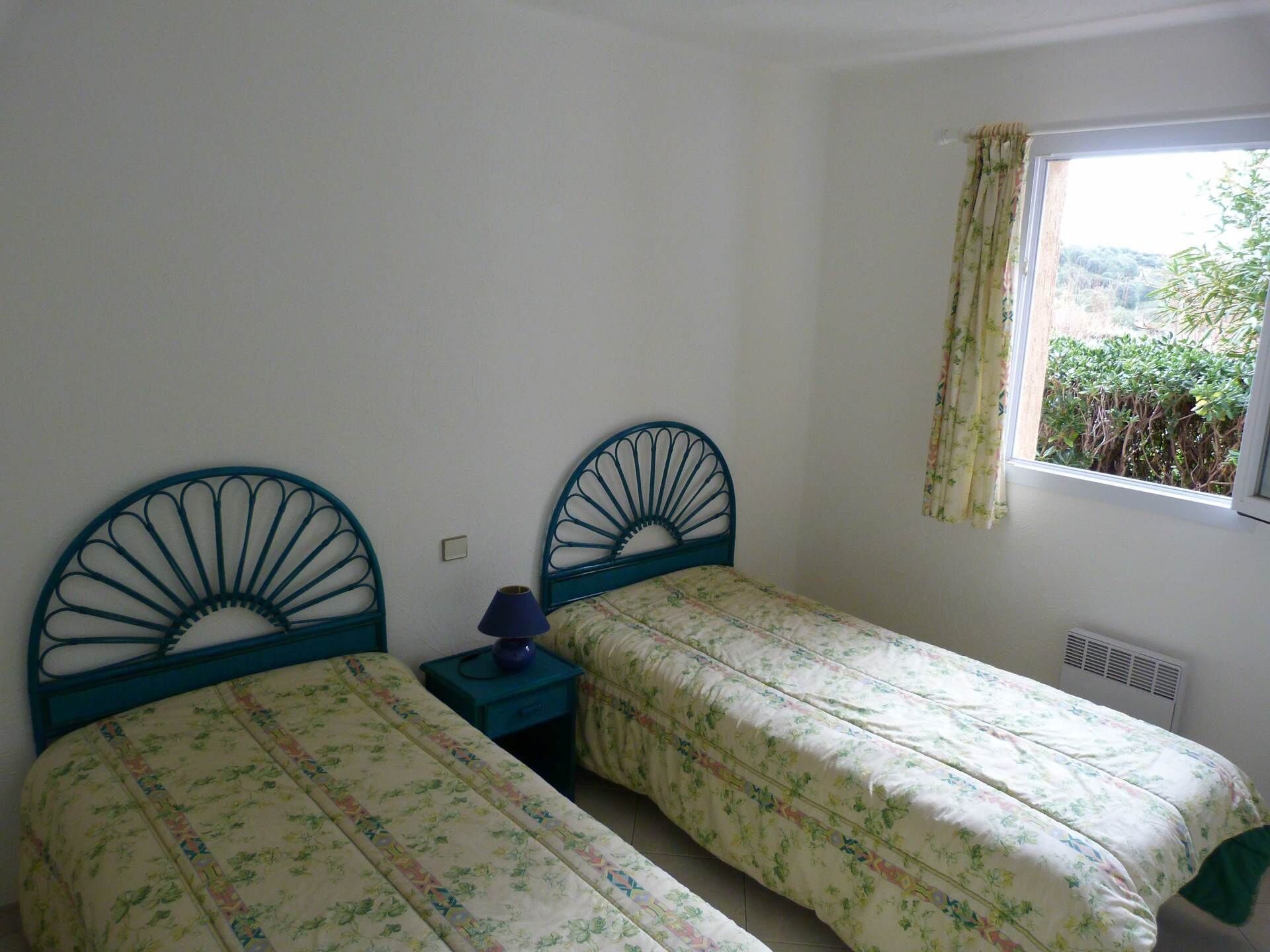 A bedroom with two single beds, a nightstand, a window with curtains, and a view of greenery outside.
