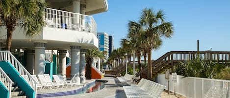 A resort with a pool, lounge chairs, palm trees, and a staircase leading to a balcony.