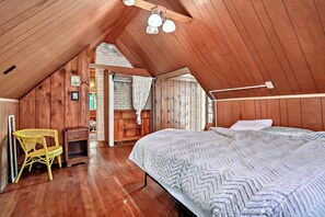Une chambre mansardée chaleureuse avec des murs en bois, un lit recouvert d’une couverture à motifs, une chaise et un luminaire suspendu.