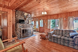 L’intérieur d’un chalet en bois, avec une cheminée en pierre, un canapé, un téléviseur et un lustre.