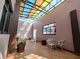 Un vaste espace intérieur doté d’un haut plafond, d’une toiture en verre et offrant une vue sur le ciel. On y trouve plusieurs chaises et tables disposées pour accueillir les visiteurs.