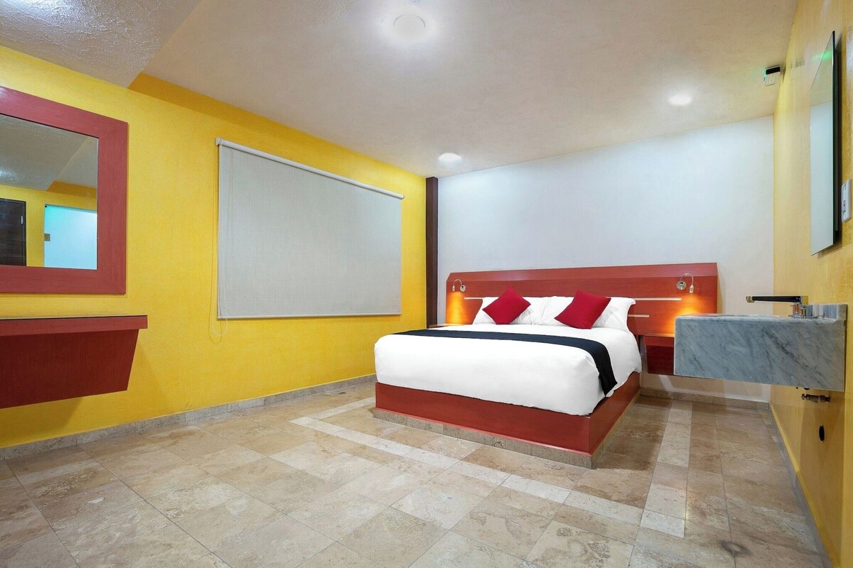 A hotel room with a bed, a bedside table, a mirror, a wall-mounted TV, and a bathroom with a sink and mirror.