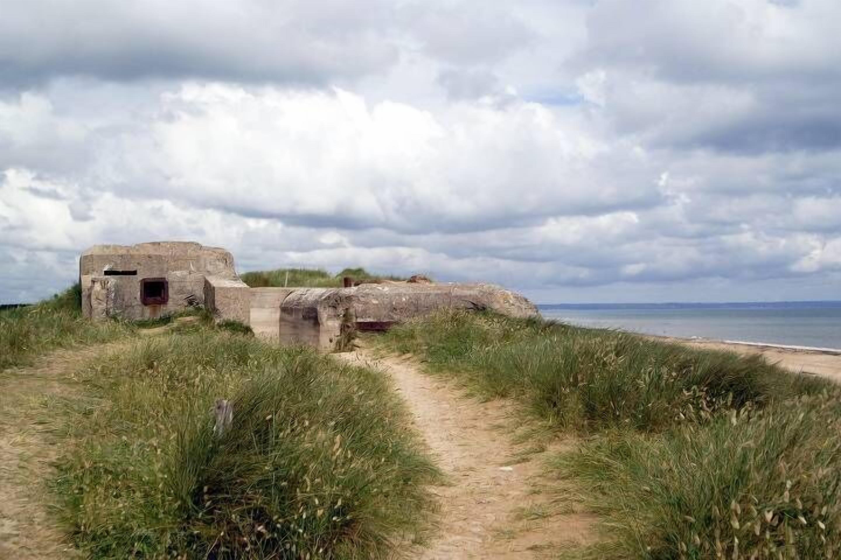 Un sentier côtier mène à une structure en pierre dotée d’une petite ouverture, entourée de dunes herbeuses et avec une plage en arrière-plan.
