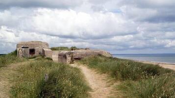 Un sentier côtier mène à une structure en pierre dotée d’une petite ouverture, entourée de dunes herbeuses et avec une plage en arrière-plan.