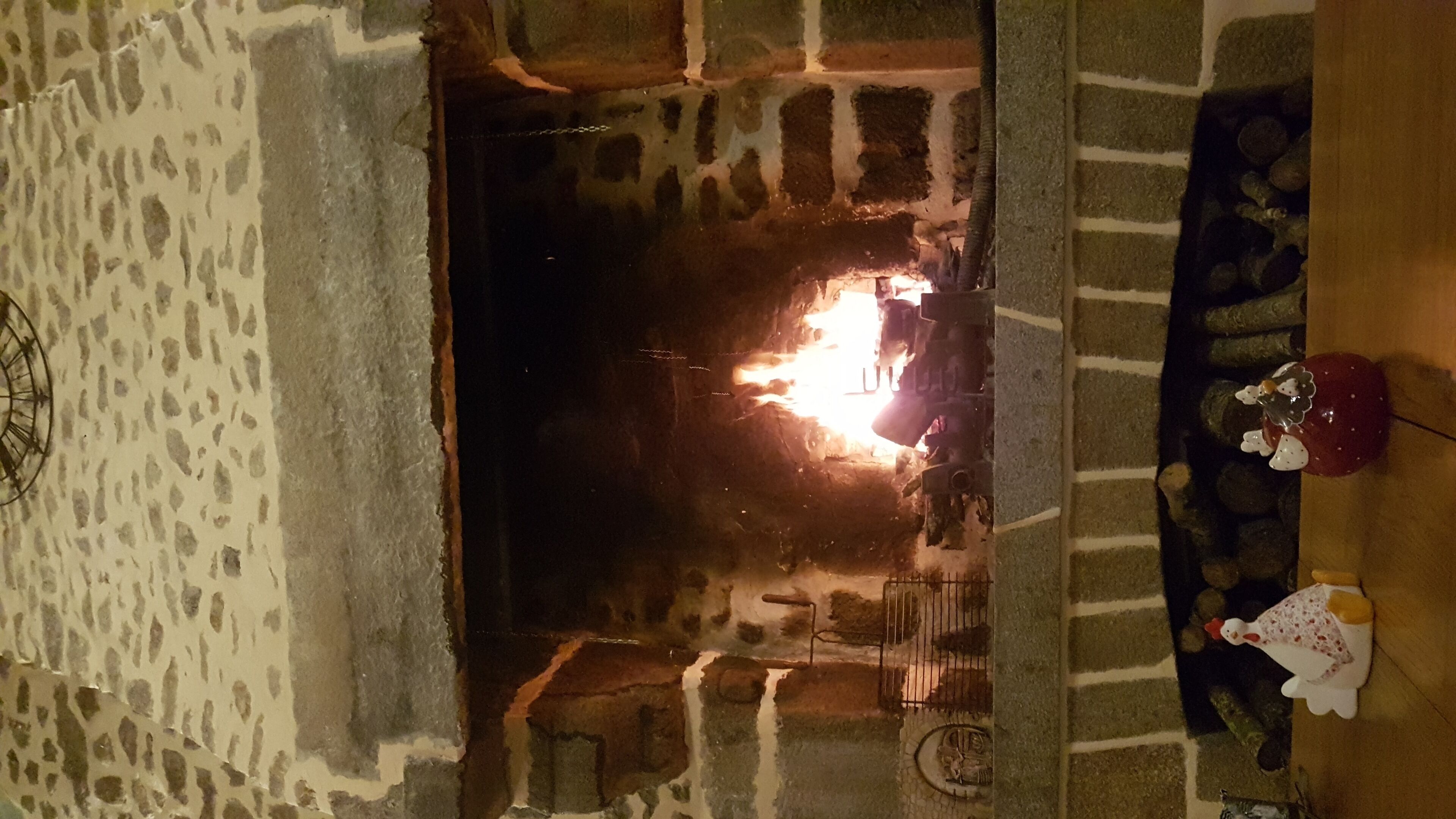 Una chimenea de piedra con fuego encendido, un rústico banco de madera a la derecha y un cerámico gallo decorativo sobre el banco.