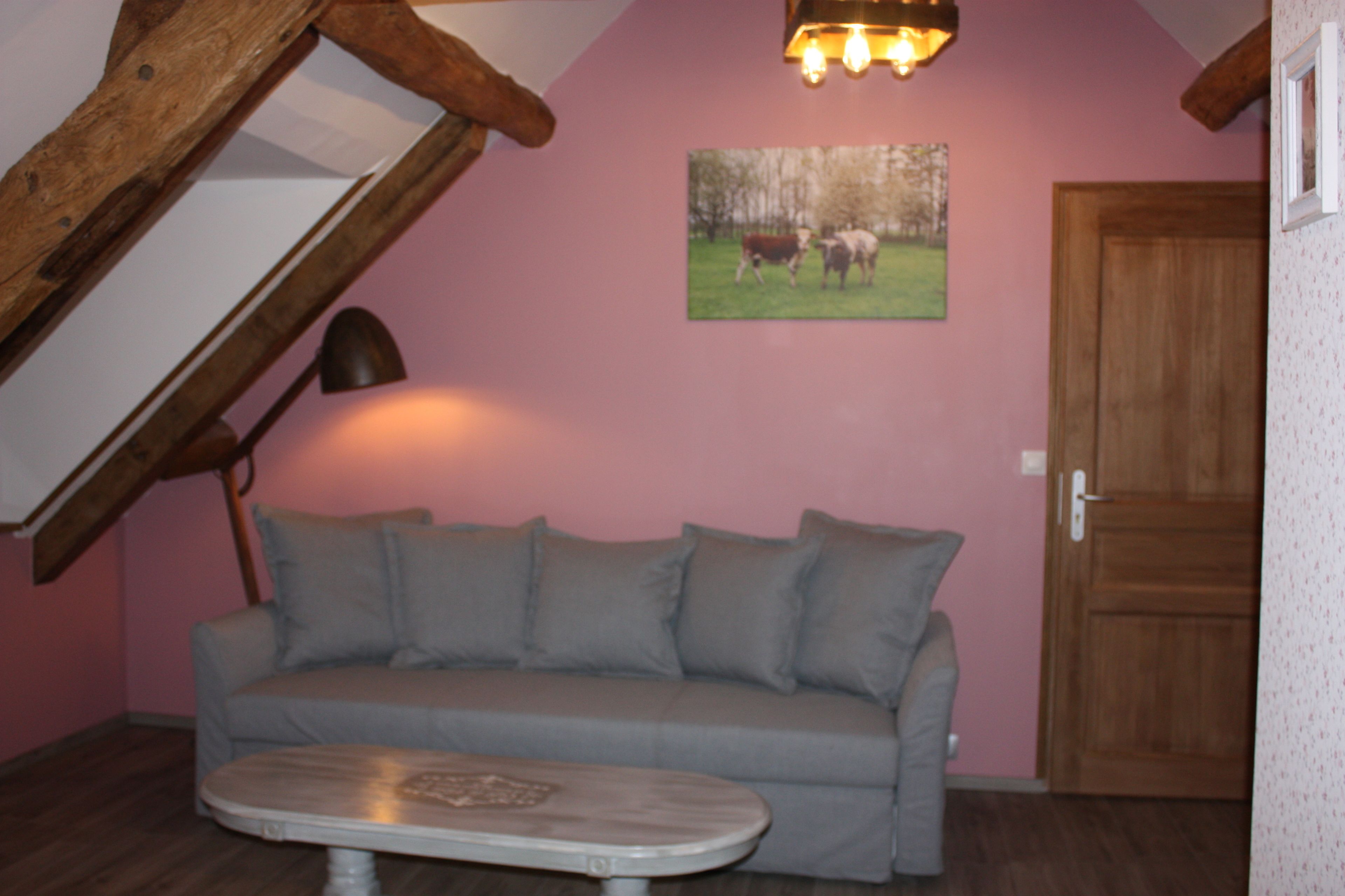 Una habitación con un sofá gris, una mesita, una viga de techo de madera, una puerta y un cuadro colgado en la pared con vacas.
