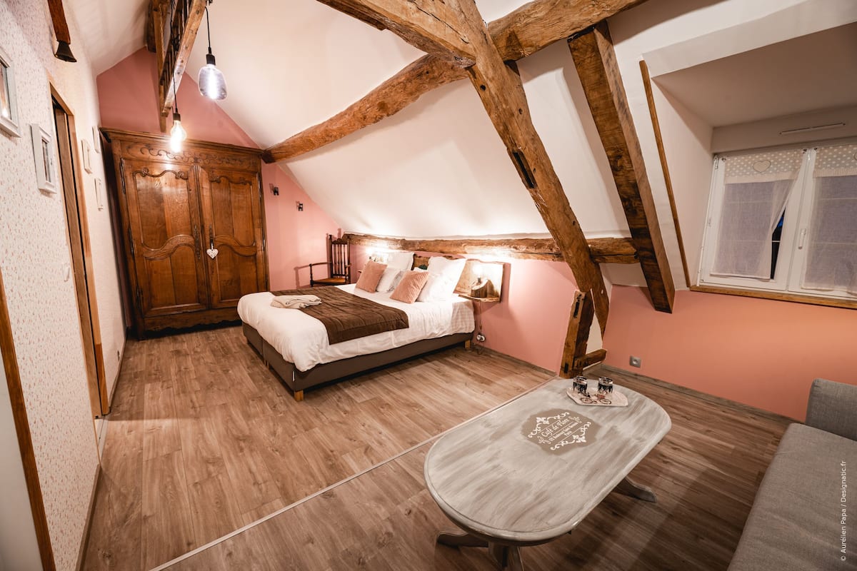 Un dormitorio acogedor con techo de madera, una cama con almohadas, una mesa con mantel individual y una ventana con cortinas.