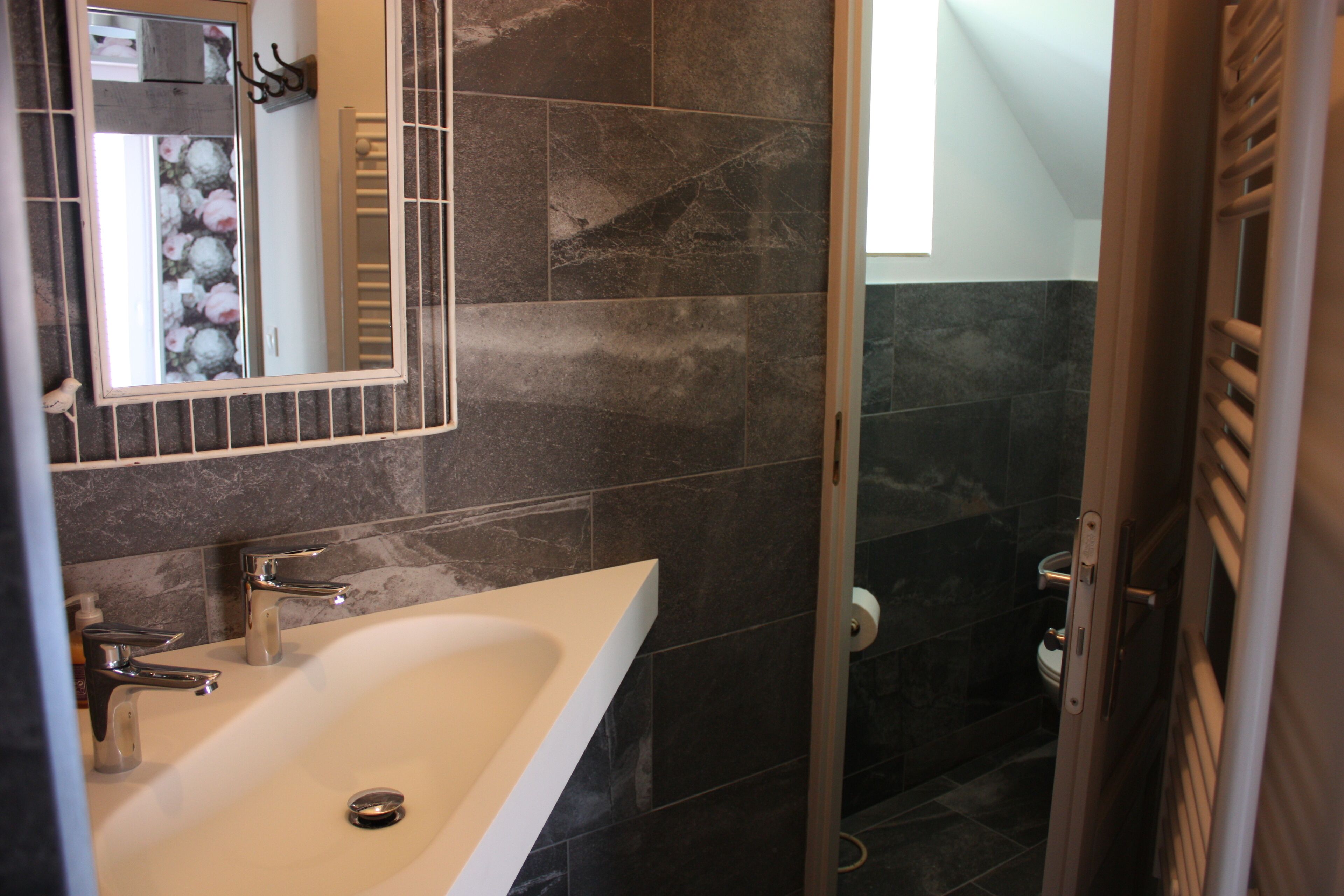 A bathroom with a white sink, two chrome faucets, a mirror with a floral frame, and dark tiled walls.